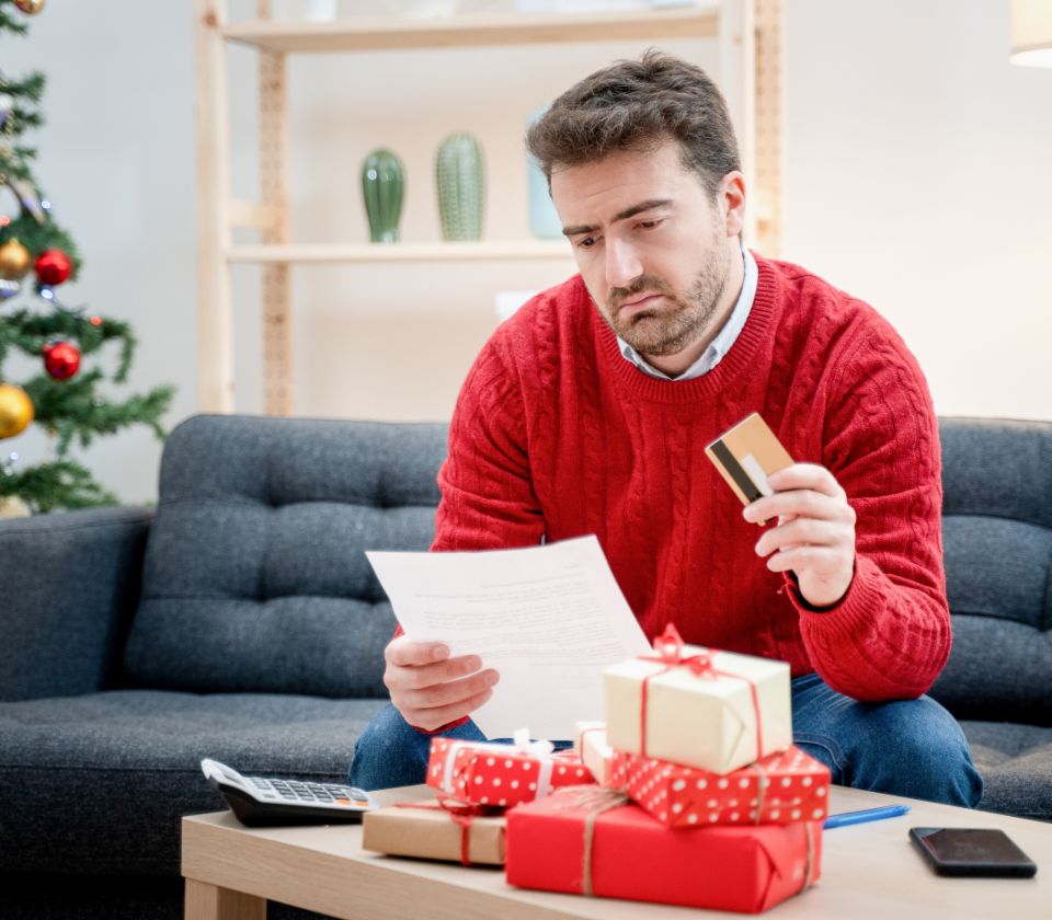 Men in a christmas decorated room sitting on the couch. He holds an invoice and creditcard in his hands and looks critical. 