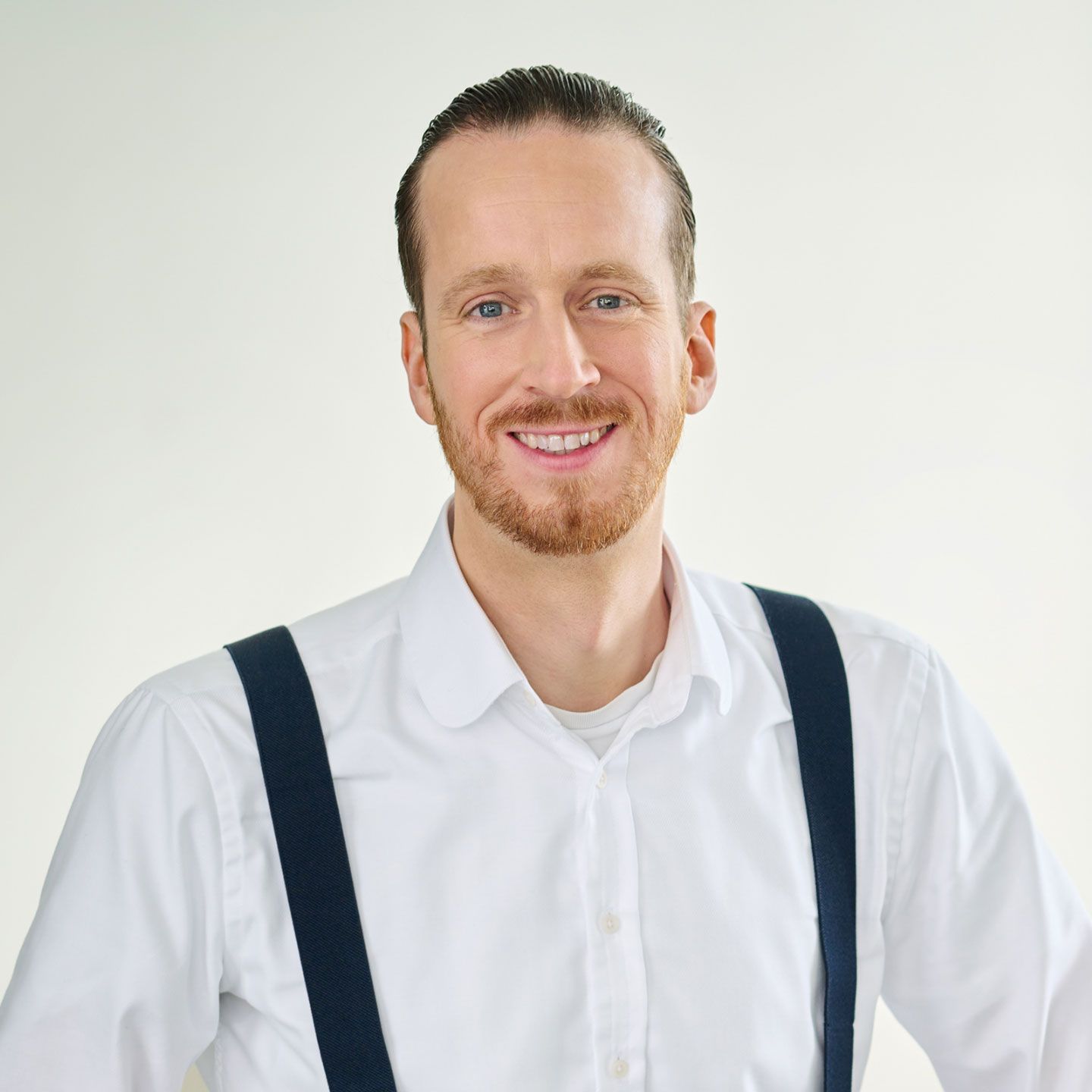 Sebastian Pollmer, muž v bielej košeli a modrých trakoch stojaci vo svetlej miestnosti.