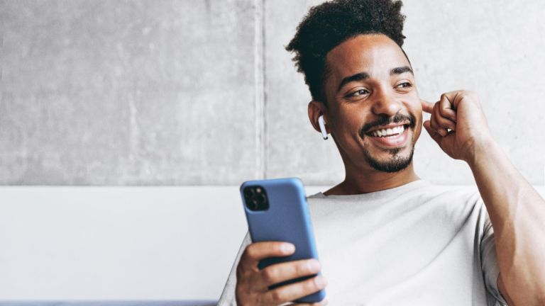 Young fun smiling african american man in beige t-shirt sitting on grey sofa indoors apartment use air pods mobile cel phone listen to music podcast looking aside resting on weekends staying at home