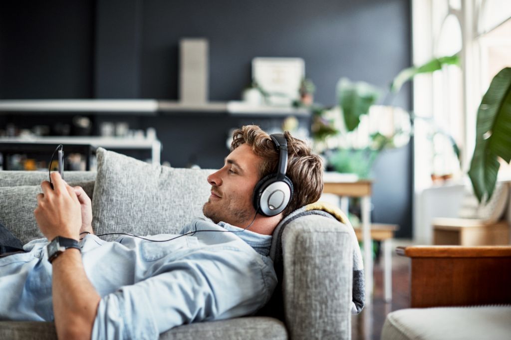 Mladý muž leží na gauči vo svojej obývačke, má na sebe slúchadlá a s úsmevom sa pozerá na svoj smartfón, zatiaľ čo si užíva hudbu alebo podcast.