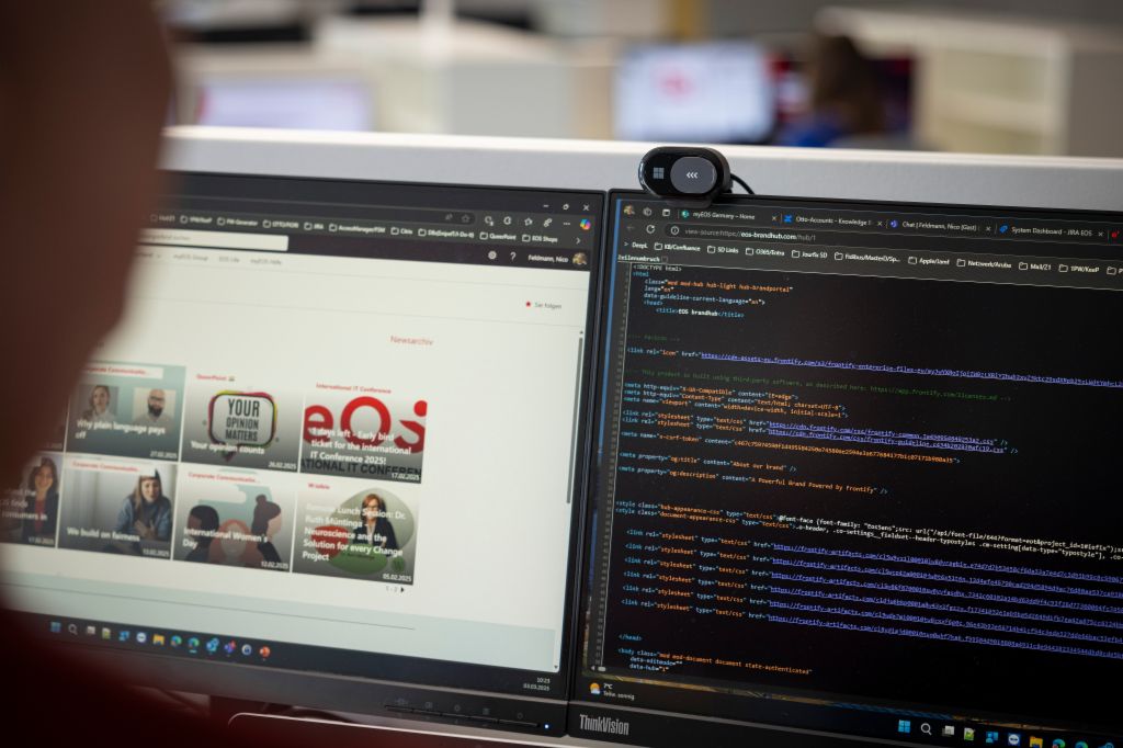 An over-the-shoulder view of a person working at a desk with two computer monitors. The left screen displays a webpage featuring various articles, including "YOUR OPINION MATTERS" and "International Women's Day." The right screen shows lines of programming code with syntax highlighting. A webcam is mounted on top of the right monitor, suggesting a typical office or developer workspace.