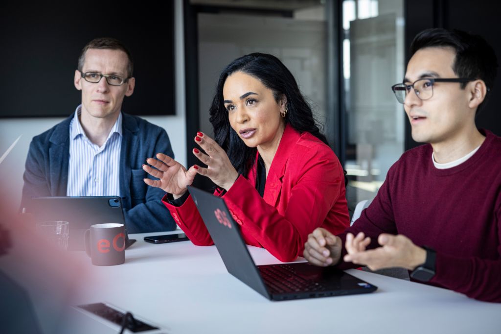 Three professionals are seated around a modern conference table, engaged in a discussion. A woman in a vibrant red blazer is at the center, speaking animatedly with expressive hand gestures. To her left, a man in a blue blazer and striped shirt listens intently, while to her right, another man in a burgundy sweater and glasses appears to be listening or responding. A laptop is open on the table in front of the woman, and a mug with "eQ" branding is visible, suggesting a business or team meeting in a contemporary office setting.