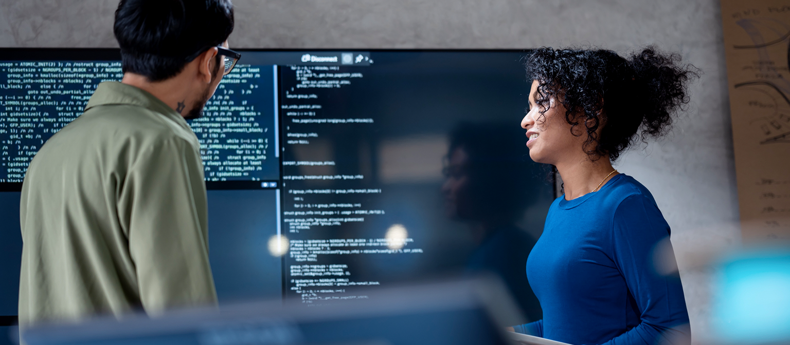 AI v inkase: Na tomto obrázku dvaja profesionáli spoločne analyzujú programový kód pre automatizované procesy na veľkom displeji. Scéna ilustruje praktickú aplikáciu umelej inteligencie (AI) a robotizovanej automatizácie procesov (RPA) v správe dlhov – príklad digitálnej transformácie v správe pohľadávok.