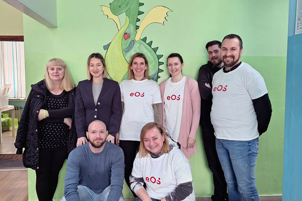 Osem ľudí stojí pred stenou, na ktorej je namaľovaný zelený drak. Osoby sú usporiadané do dvoch radov: dvaja čupia vpredu a šiesti stoja vzadu. Štyri osoby majú na sebe biele tričká s červeným logom EOS. Skupina sa usmieva do fotoaparátu. Na podlahe vpredu vpravo vidíme niekoľko fliaš a kartónov od nápojov.