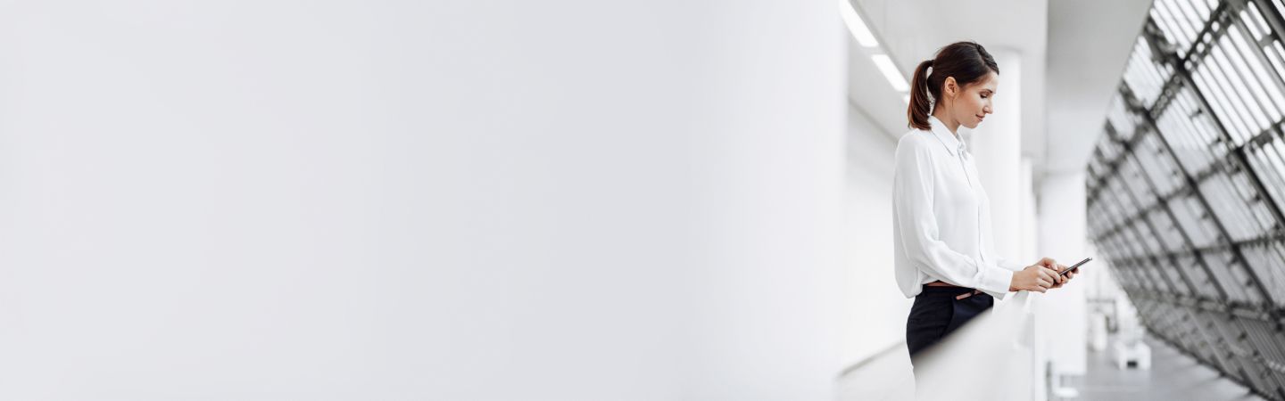 Section Stage Company Young woman in white blouse stands on a gallery in front of a high glass façade and looks at her smartphone.