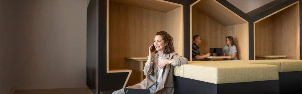 Usmievavá osoba sedí v modernej kancelárskej oddychovej zóne, telefonuje a pracuje na notebooku. V pozadí pracujú ďalší dvaja ľudia.