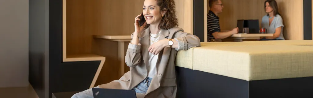 Usmievavá osoba sedí v modernej kancelárskej oddychovej zóne, telefonuje a pracuje na notebooku. V pozadí pracujú ďalší dvaja ľudia.