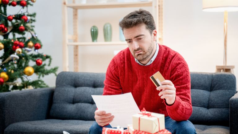 Men in a christmas decorated room sitting on the couch. He holds an invoice and creditcard in his hands and looks critical. 