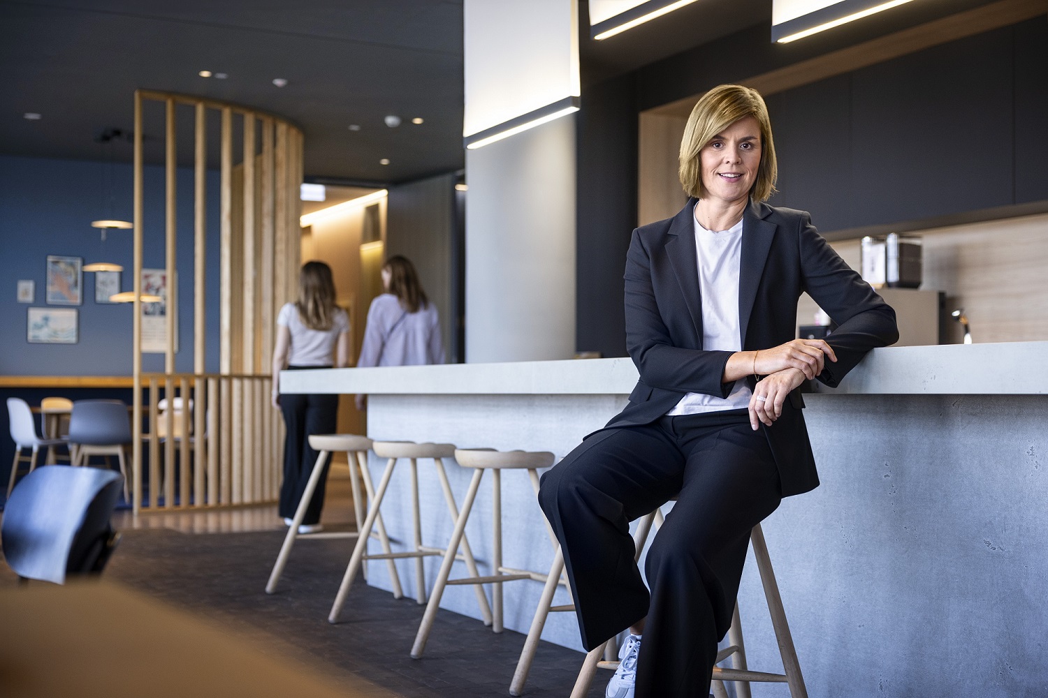 Dr. Eva Griewel, finančná riaditeľka (CFO) skupiny EOS, sedí v spoločenskom priestore na vysokom stoličke, oblečená v čiernom bleyzeri.